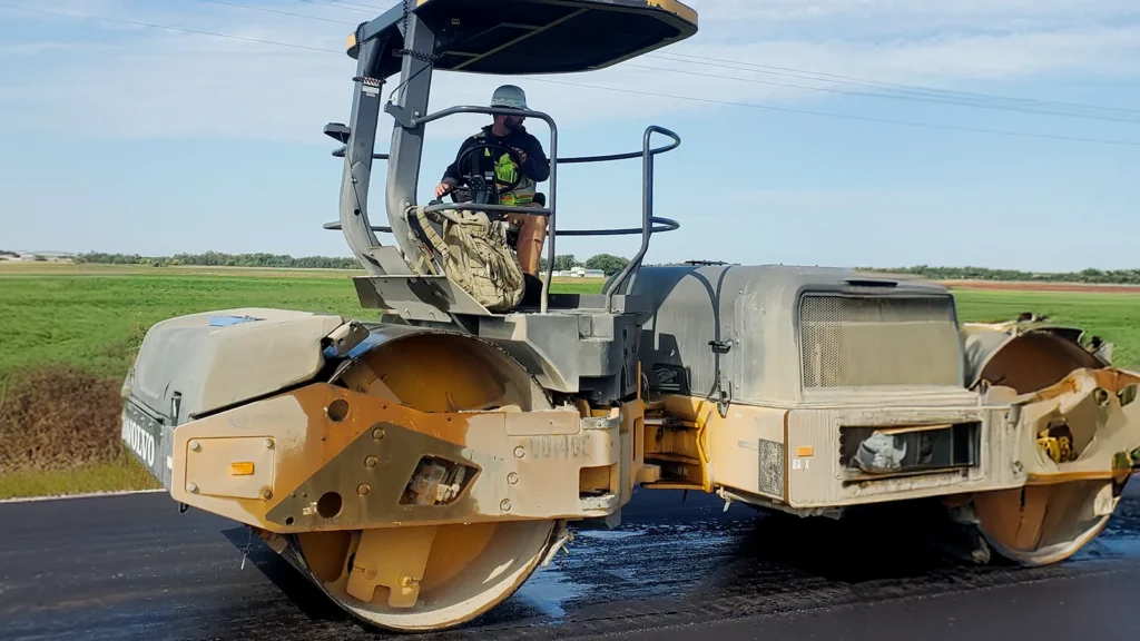 A worker operating a steam roller over a freshly asphalted roadway